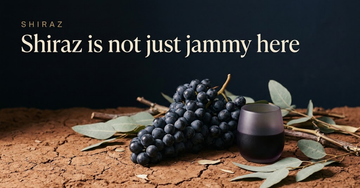 A dense cluster of dark Shiraz grapes rests on cracked red earth beside a small glass of deep purple wine — Shiraz is not just jammy here