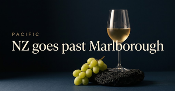A glass of pale gold wine sits on black volcanic stone beside a small cluster of green grapes in soft coastal light — NZ goes past Marlborough