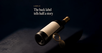 An unlabelled bottle lies on its side, back facing up with a blank paper label and a loupe propped against it — The back label tells half a story