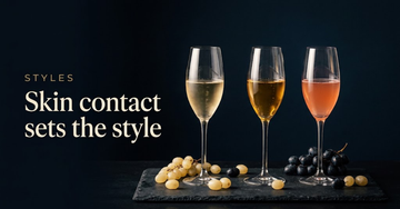 Three champagne glasses holding pale gold, deep gold and salmon pink wine, white and dark grapes scattered on dark slate — Skin contact sets the style