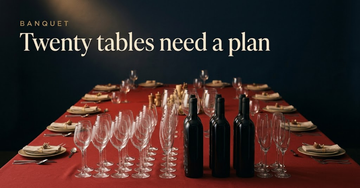 Rows of wine glasses and unopened bottles fanned across a long red banquet cloth, round table settings receding behind — Twenty tables need a plan