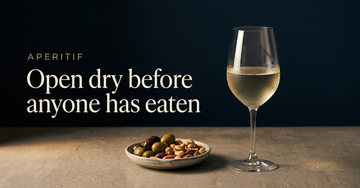A single chilled glass of pale wine beside a small dish of olives and nuts on a bare counter, no plates laid yet — Open dry before anyone has eaten