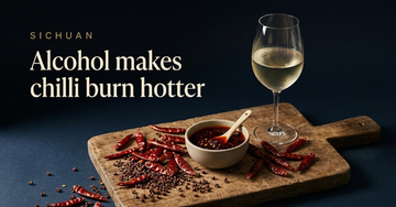 Dried red chillies and whole Sichuan peppercorns scattered across a wooden chopping board beside a bowl of chilli oil — Alcohol makes chilli burn hotter
