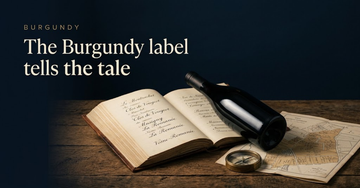 An unlabelled Burgundy bottle leaning against a ledger open to handwritten vineyard names, a brass compass and folded map on oak — The Burgundy label tells the tale