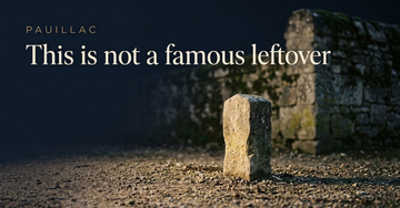 A modest stone marker standing alone in gravel soil, a larger shadowed estate wall out of focus beyond it — This is not a famous leftover