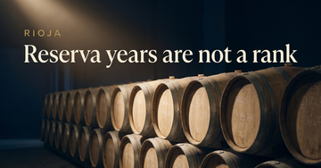 Stacked American oak barrels lining a dim bodega corridor, chalk vintage marks faint on the barrel ends — Reserva years are not a rank