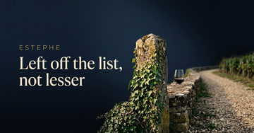 A weathered stone boundary marker half covered in ivy at a vineyard edge with a gravel path beyond — Left off the list, not lesser